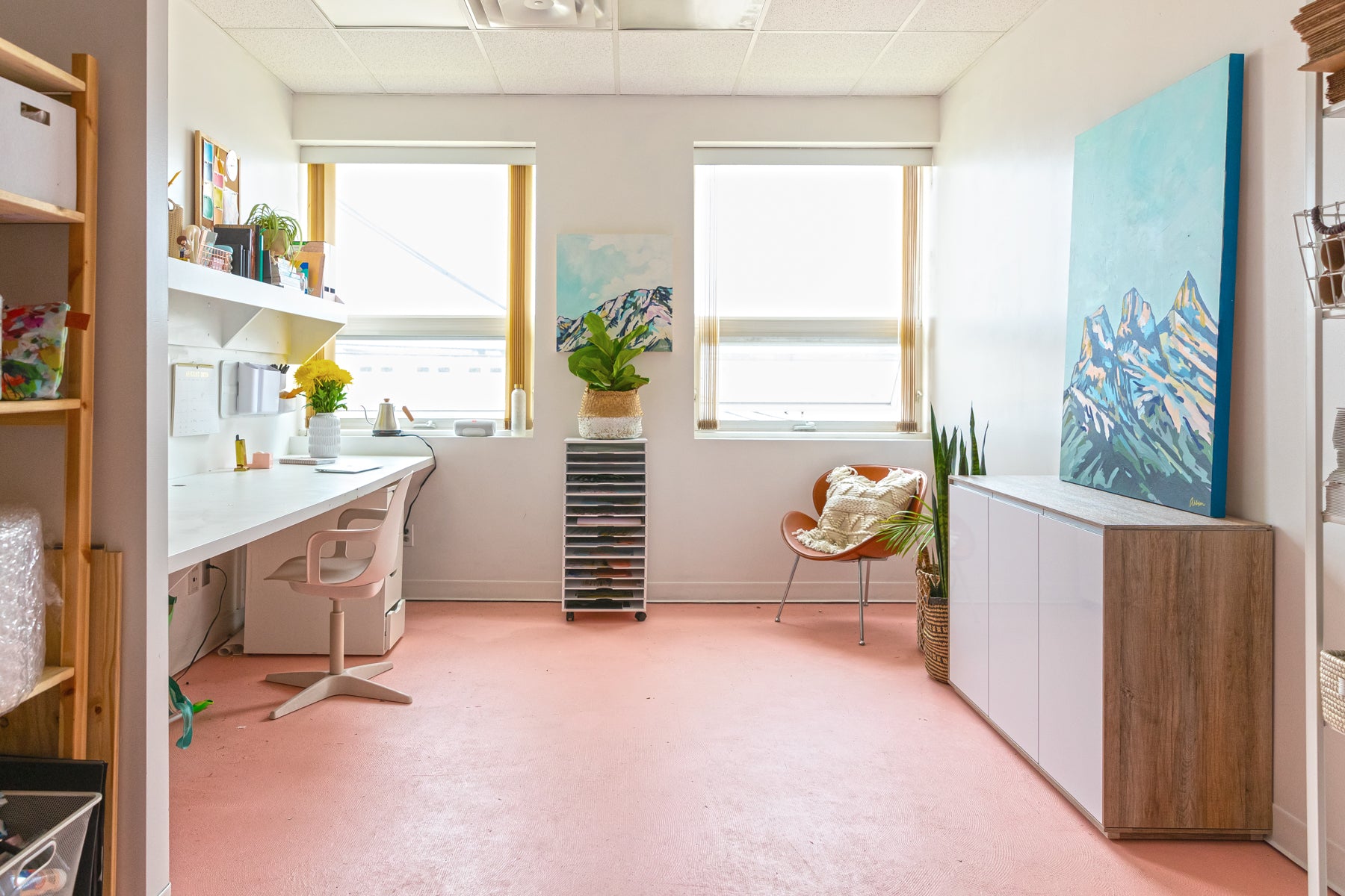 Amy Dixon's studio in Edmonton, Alberta, Canada. It has pink floors with a clean, white desk on the left side, two large windows on the far wall, and a cabinet on the right side. Her mountain painting Three Sisters I is positioned on top of the cabinet.