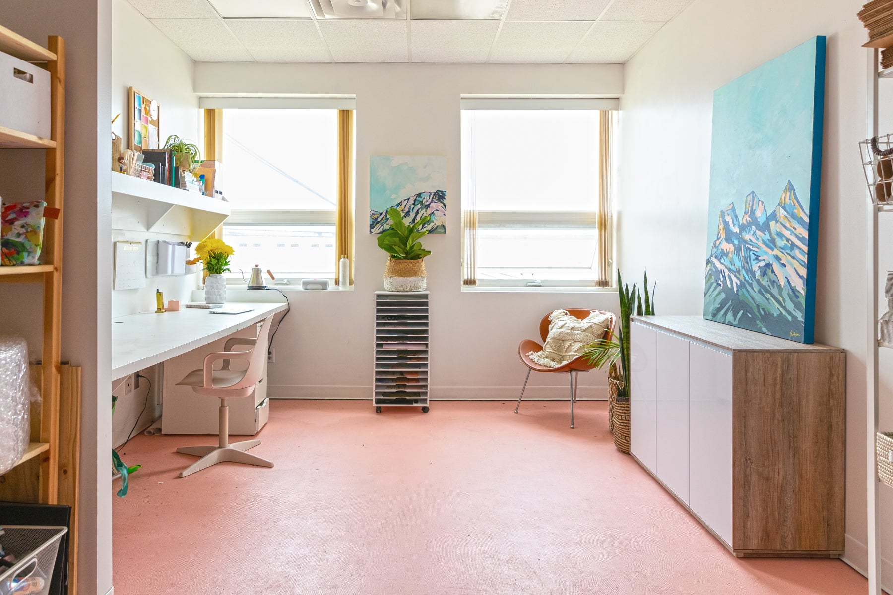 Amy Dixon's studio in Edmonton, Alberta, Canada. It has pink floors with a clean, white desk on the left side, two large windows on the far wall, and a cabinet on the right side. Her mountain painting Three Sisters I is positioned on top of the cabinet. 